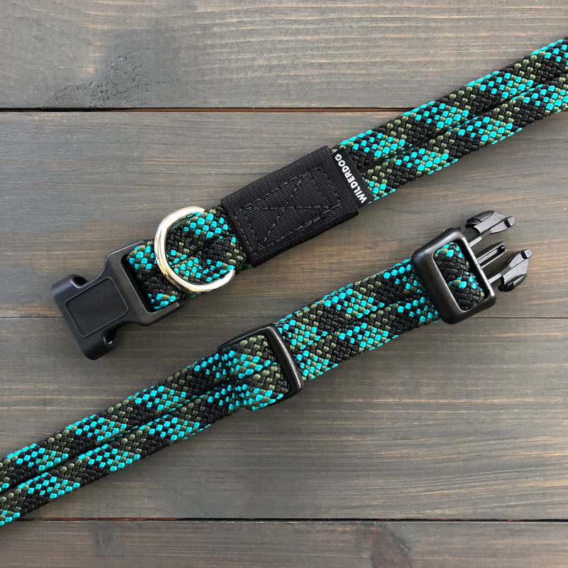 A black, blue, and green woven Granite Collar laying flat on a wooden surface. This durable dog collar features a plastic buckle, heavy-duty hardware with a metal D-ring, and a tag with "Wilderdog" printed on it. Designed for longevity, the collar has an adjustable strap for sizing flexibility.